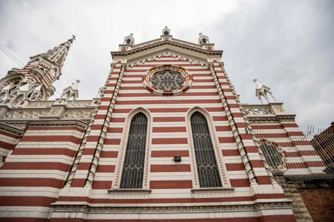 Santuario Nuestra Señora del Carmen en primerísimo primer plano de fachada gótica vista desde abajo sobresaliendo las franjas horizontales rojas y blancas, los adornos del techo y la aguja de la torre principal. El cielo es gris.