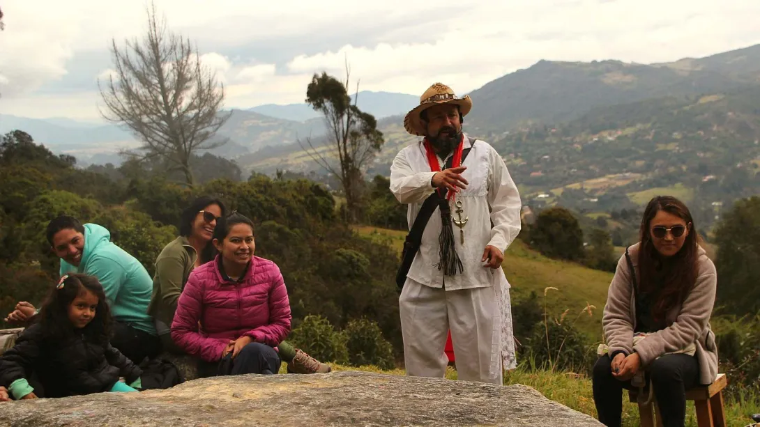  Grupo de personas, con ellos una niña, en el campo, rodeados por montañas. Un hombre vestido como campesino está hablando. 