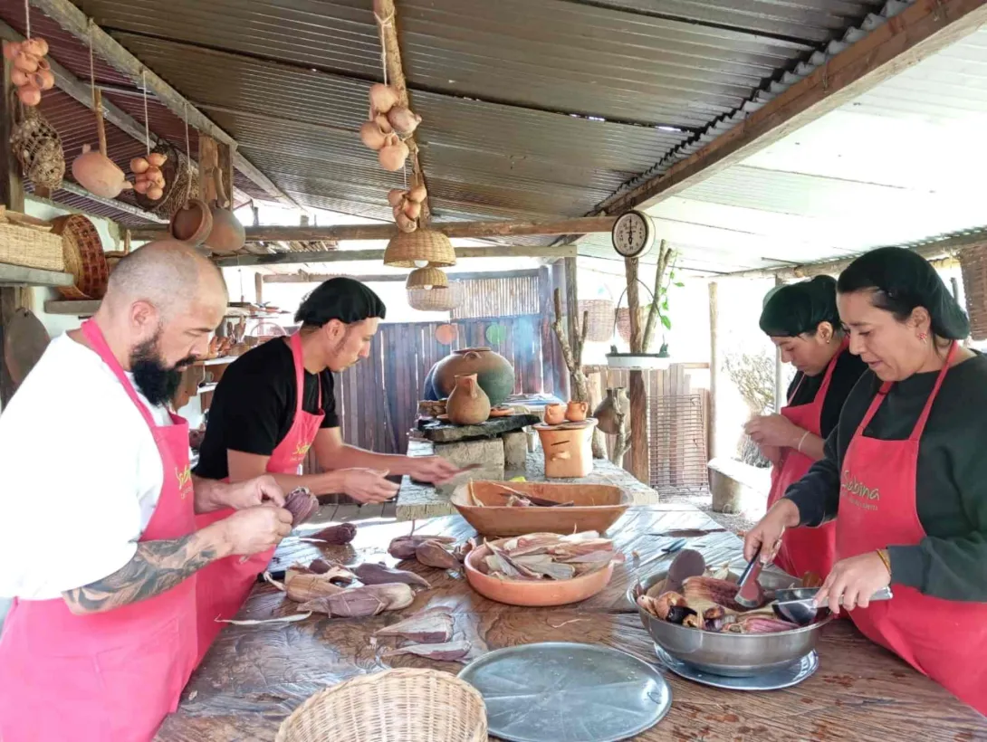 Dos hombres y dos mujeres con delantales rojos, cocinando mazorcas en una cocina artesanal.