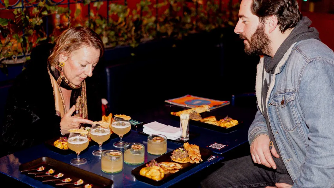 Mujer adulta rubia y jóven rubio sentados en un restaurante alrededor de una mesa con copas y vasos con licor y con sendos platos repletos de comida criolla colombiana como empanadas, papas y arepas.