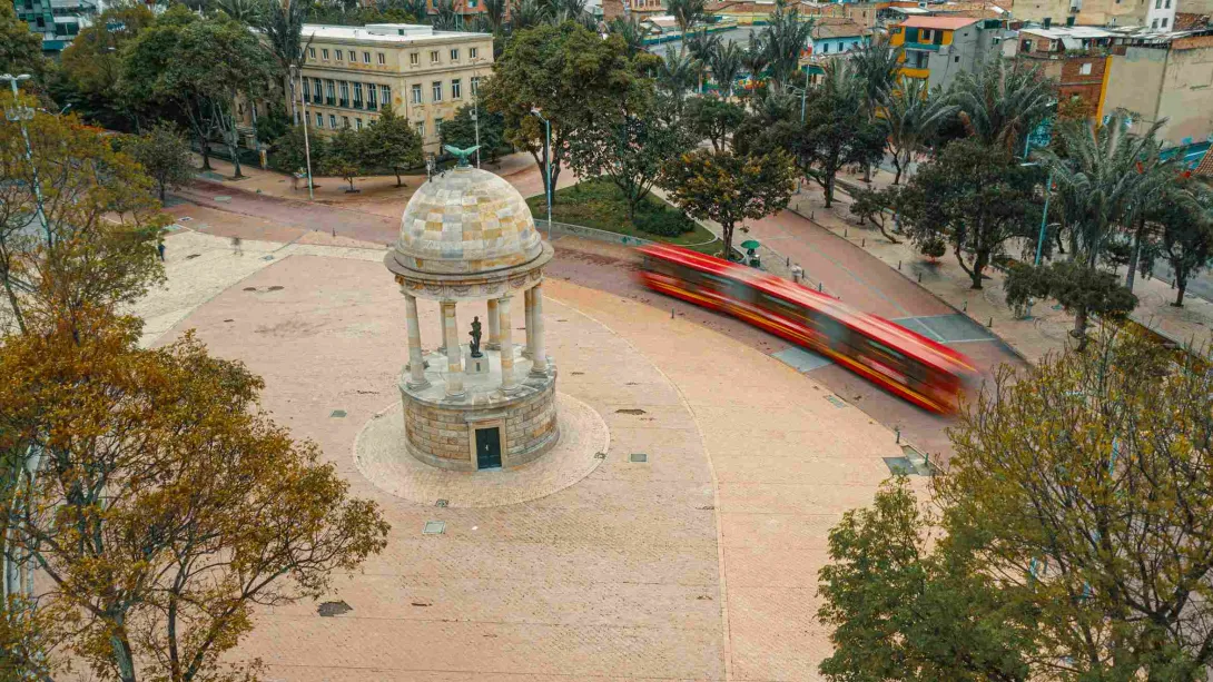 El Parque de los Periodistas visto desde arriba, rodeado por árboles frondosos. Un TransMilenio deja la estela de su paso a gran velocidad.