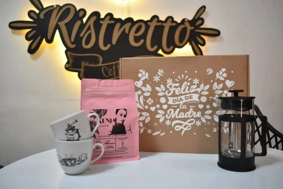 Una mesa blanca sobre la que estan dos pocillos, un paquete de café, una máquina para prepara café, una caja con un aviso que dice "feliz día de la madre" y al fondo sobre la pared el aviso que dice Ristretto.