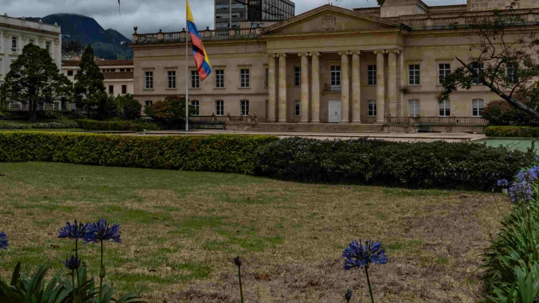 Fachada de la Casa de Nariño, edificio neo colonial en baldoza amarilla en el que se ven cuatro columnas. Al frente hay un amplio jardín con vegetación y una bandera de Colombia gigante.