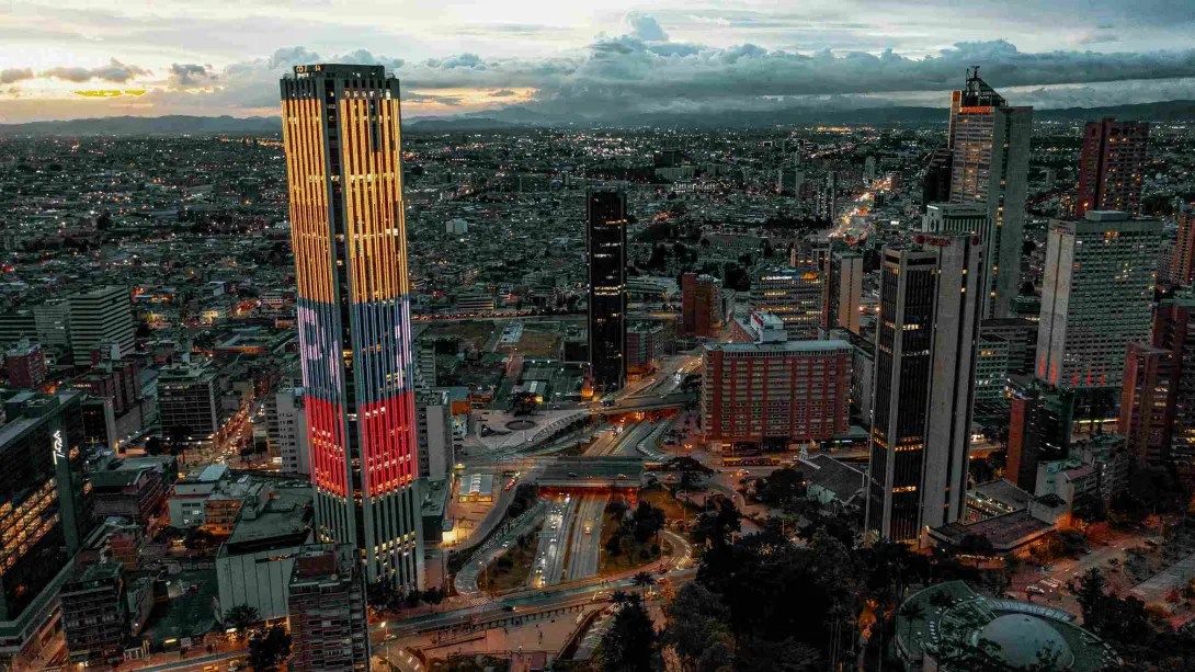 En el atradecer iluminada la Torre Colpatria con los colores de la bandera de Colombia. Al fondo las luces de la inmensa ciudad en la que destacan algunos edificios altos y el Planetario.