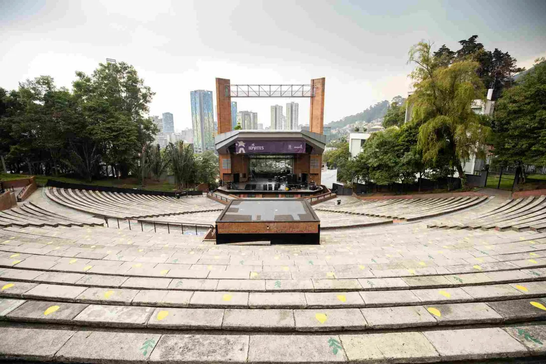 Teatro al aire libre en ladrillo, de día, se ven escalinatas hacia la tarima, que está cubierta por un techo, dos columnas en ladrillo y un aviso gigante donde se lee "Idartes". 