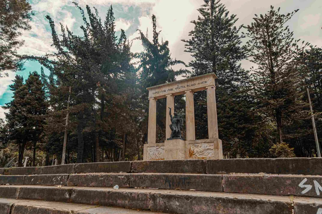 Monumento en piedra tallada con una estatua negra al frente y varios pinos altos detrás.