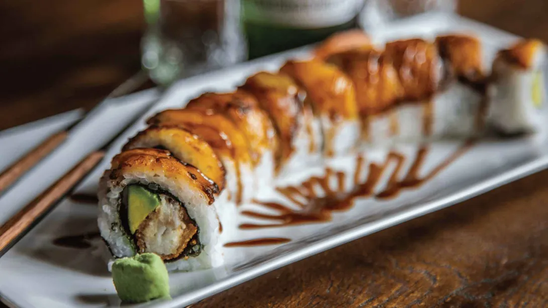 Un rollo de sushi generosamente bañado en salsa, con aguacate y un relleno crujiente, servido con wasabi en un plato blanco.