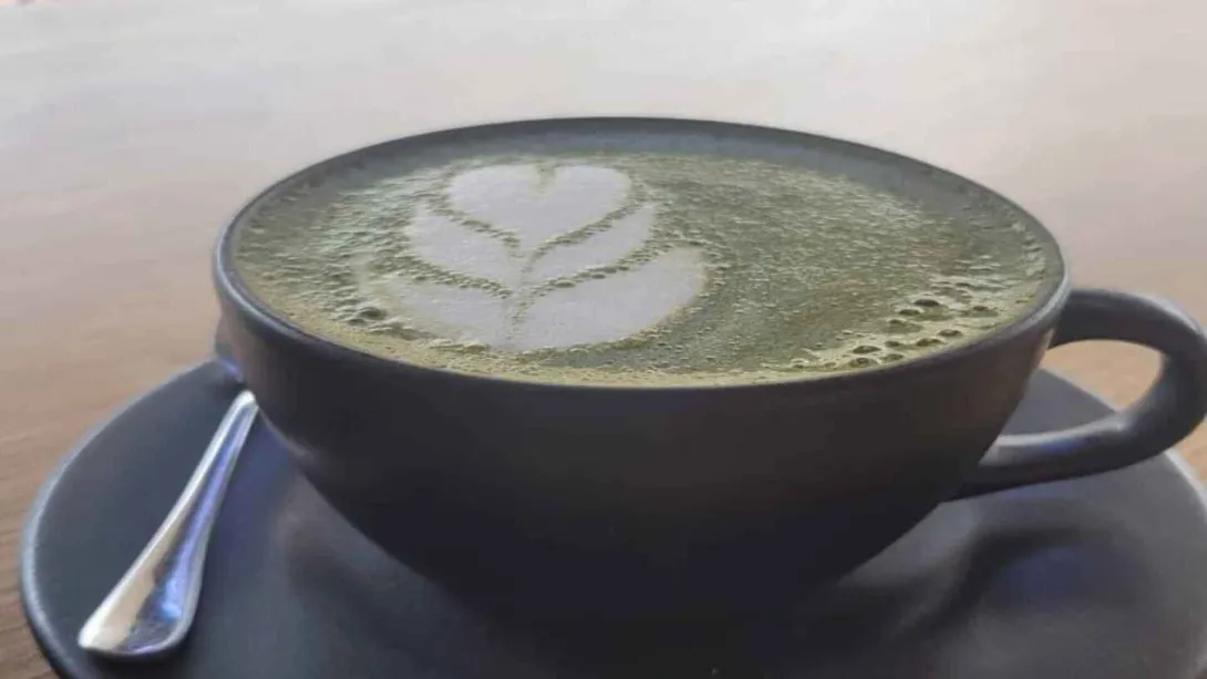  Una taza oscura de matcha latte con arte de espuma en forma de flor, sobre un platillo a juego. Pie de foto: Disfruta de la calma y el sabor único de un matcha latte perfectamente preparado.