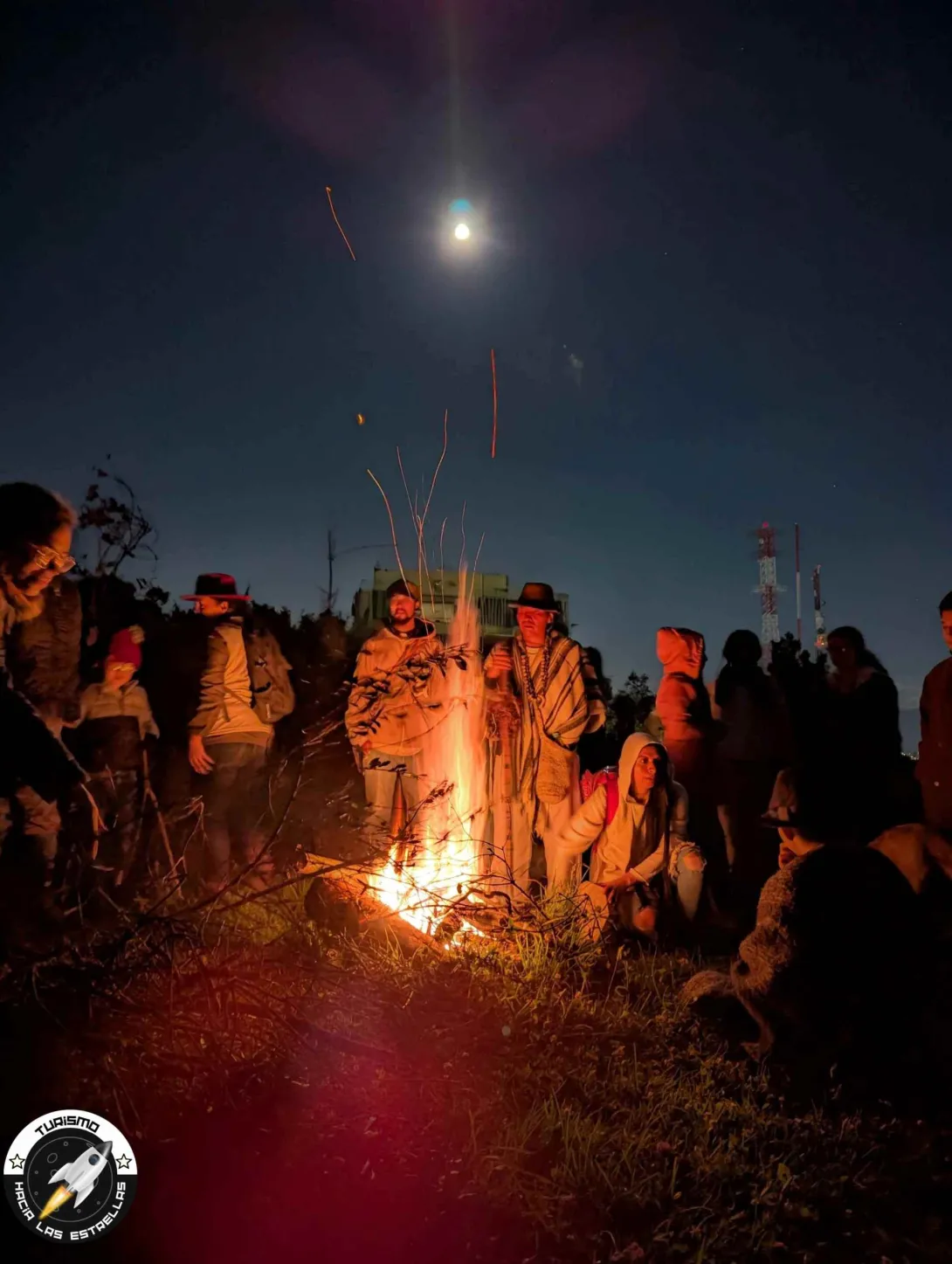 Grupo de personas entre jóvenes y adultos de noche, con chaquetas, reunidos alrededor de una chimenea. En el centro está una persona que parece un campesino o indigena con sombrero y ruana.