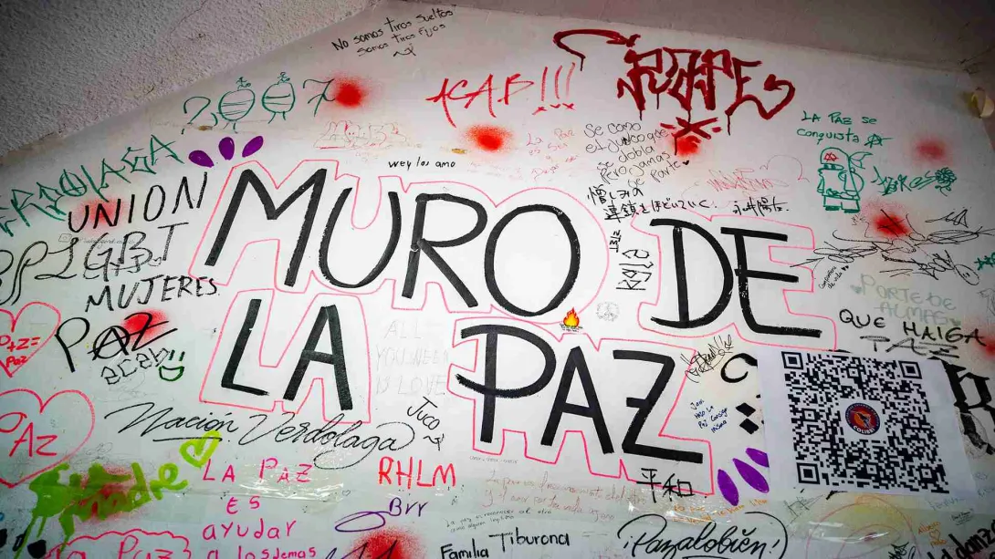 Una pared de fondo blanca llena de mensajes y nombres de personas con tintas de varios colores y al centro el aviso del nombre del lugar, con letras negras en mayúscula que dice "Muro de la Paz"