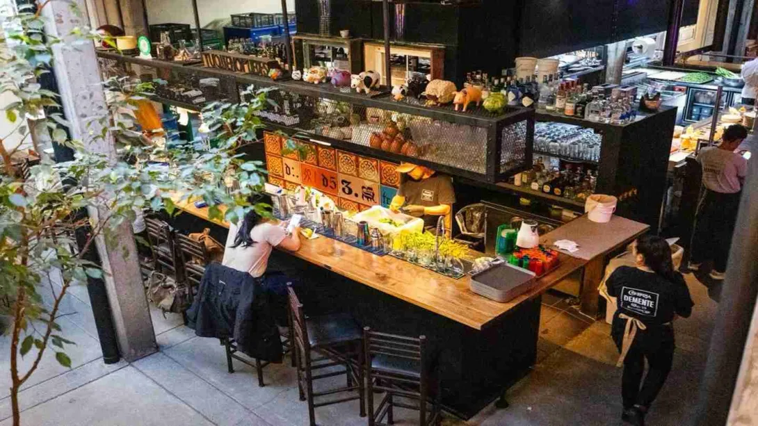 Interior local comercial en el que se ven barras para atender clientes, varias botellas de licos y dos meseros alrededor.