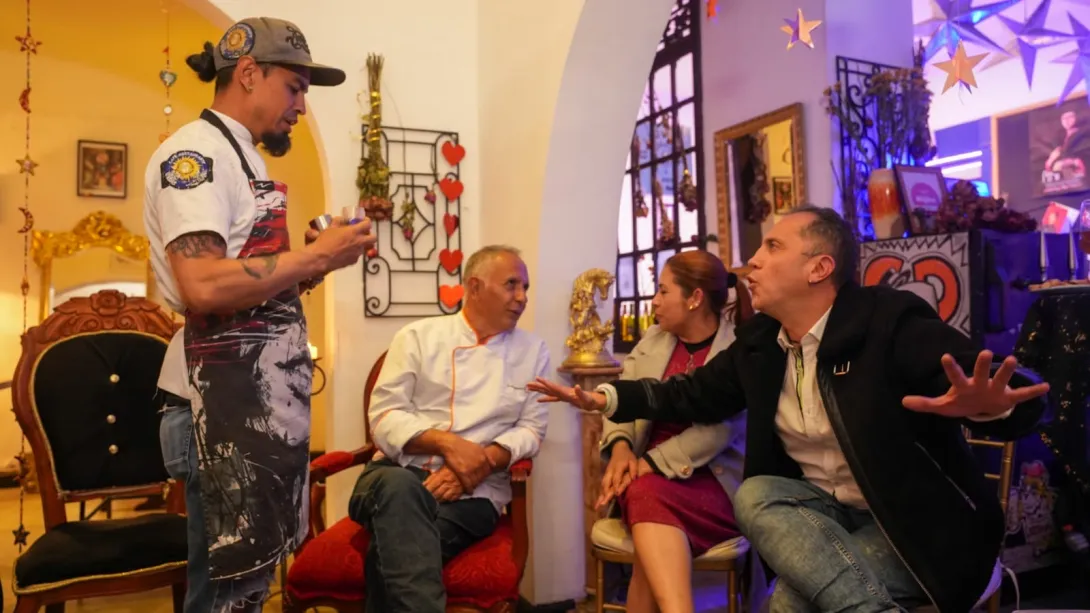 Un hombre adulto vestido como chhef y otro joven vestido con delantal conversa con una mujer y un hombre adultos. Están sentados en un salón con decoración antigua. 