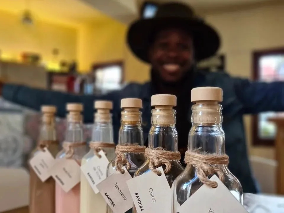  Primer plano de unas botellas de vidrio en las que resalta una leyenda que dice: "Bantura". Detrás se observa un rostro borroso de un hombre afrodescendiente con sombrero y que ríe.