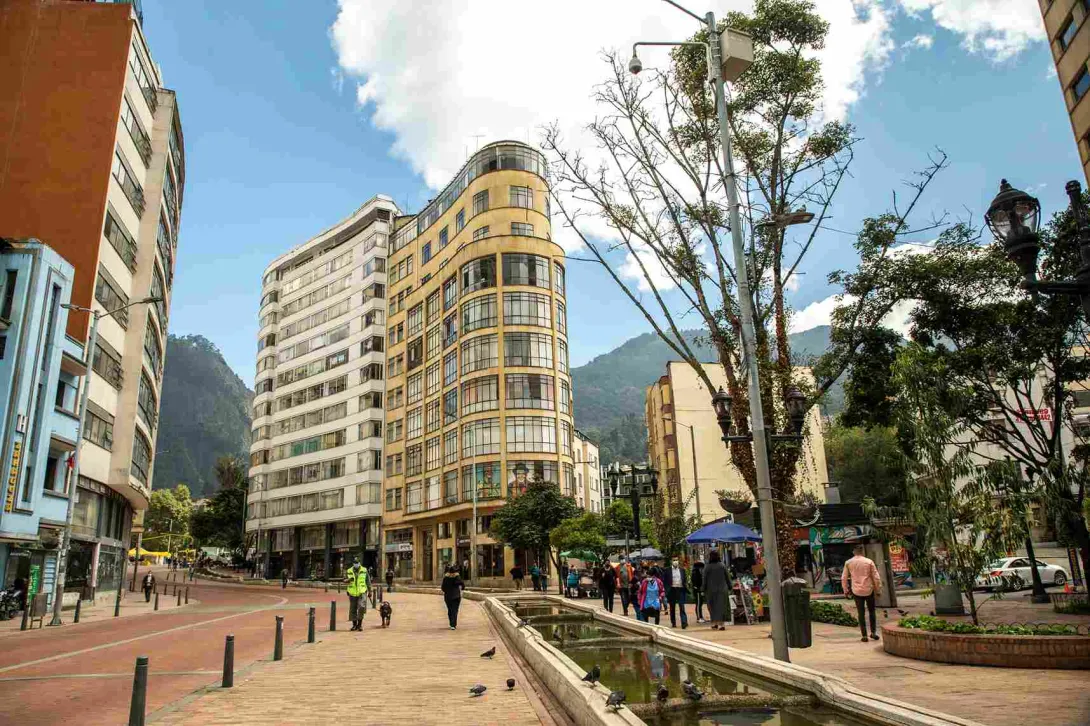 Alameda con una larga fuente de agua y en la que en una esquina hay un edificio antiguo con muchas ventanas. Hay árboles, un guarda de seguridad con un perro, y personas alrededor.
