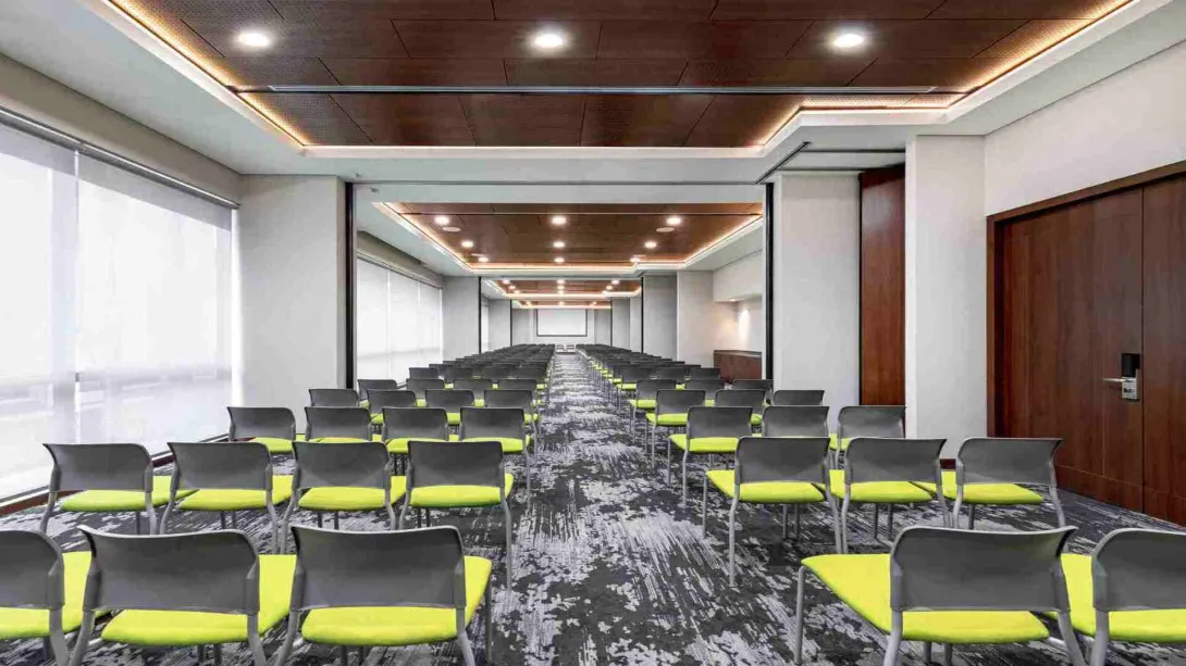 Un moderno lobby lounge con sofás verdes y grises, mesas de madera y grandes lámparas colgantes. La decoración cuenta con paneles de madera y patrones geométricos.