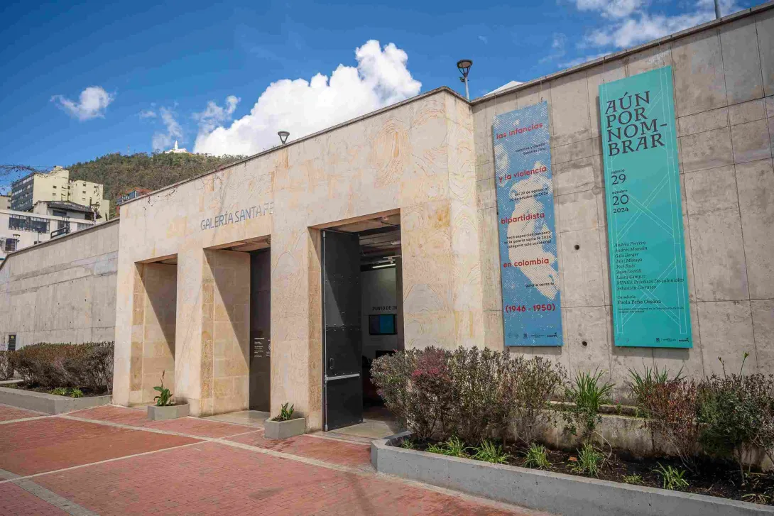 Fachada de edificación en piedra en la que destacan un par de pendones y una leyenda sobre la parte superior que dice: “GALERÍA SANTA FE”. Al fondo se observa el cerro de Monserrate. 