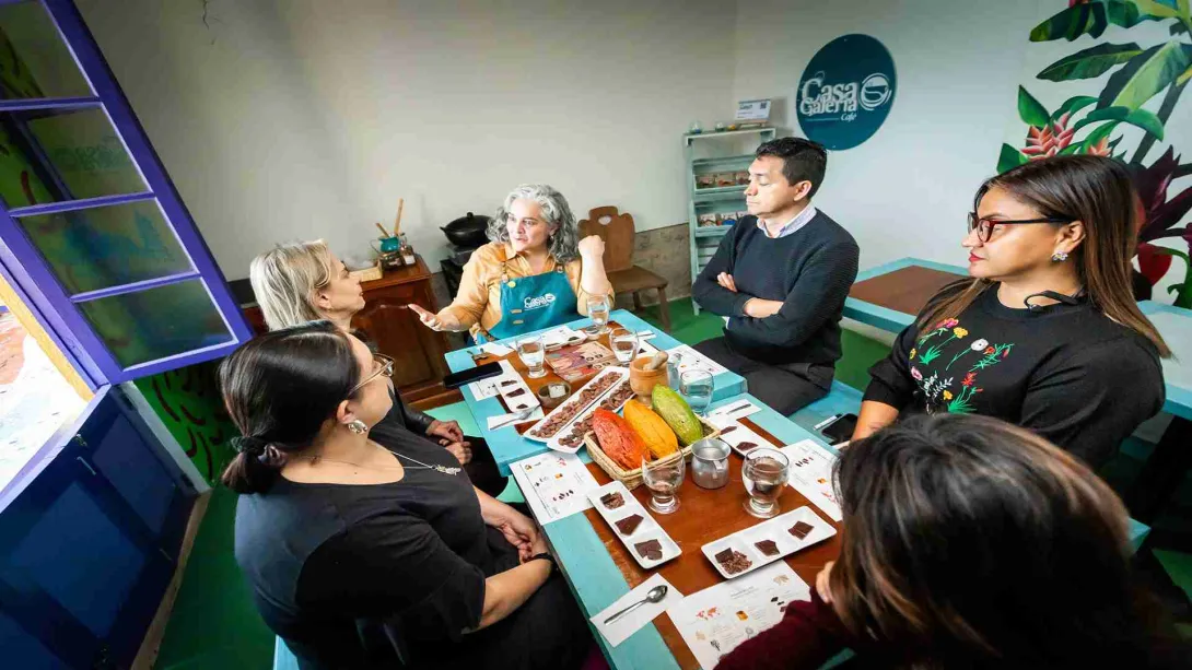 Grupo de tres mujeres y un hombre sentadas en una mesa en la que hay varias copas de cristal. Hay una mujer adulta con un delantal azul y al fondo en la pared un aviso con una leyenda que dice: "Casa Galería".