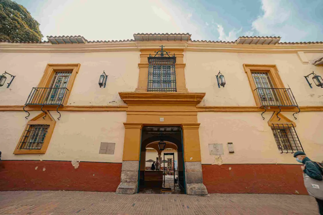 Fachada de casa antigua en la que se destacan sus colores crema, amarillo y rojo, y sus dos ventanas con rejas en hierro.