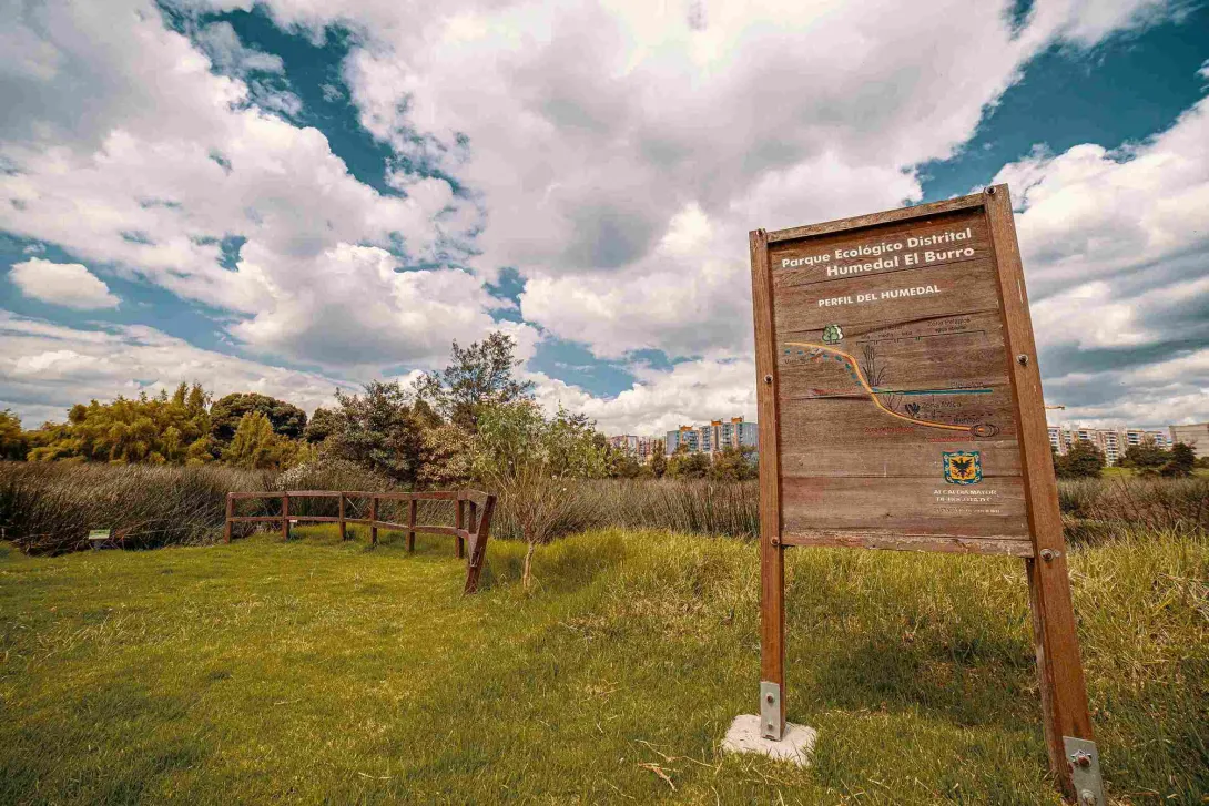 Una zona verde en la que destaca un cartel en madera con una leyenda que dice: “Parque Ecológico Distrital Humedal El Burro”.