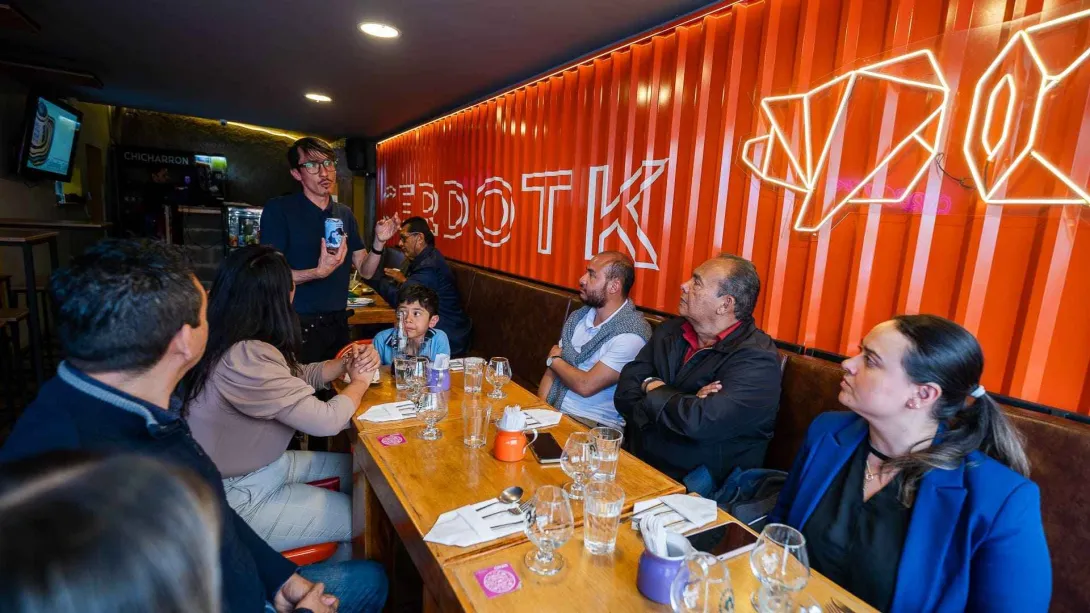 Un grupo de personas, incluido un niño, escuchando atentamente a un hombre que presenta un plato en un restaurante con decoración moderna.