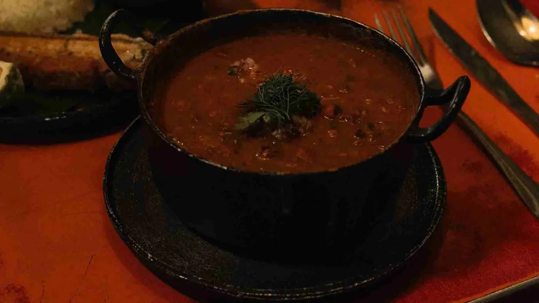  Primer plano de un guiso oscuro servido en una olla de metal rústica con hierbas, acompañado de cubiertos en una mesa.