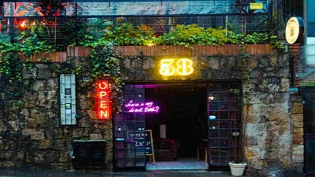  Entrada de un bar/restaurante con paredes de piedra, plantas y letreros de neón "OPEN" y "58". 