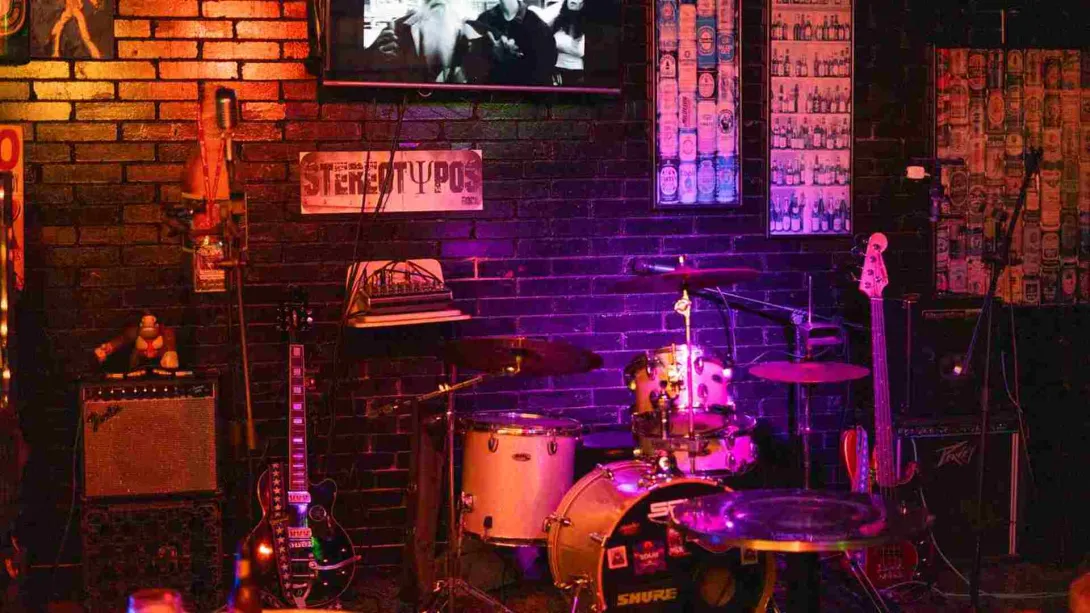 Un escenario de bar con batería, bajo y amplificadores bajo luces de colores, listo para una actuación.