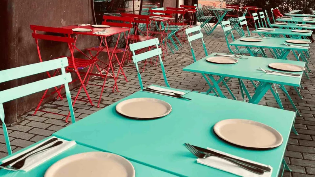 Terraza de restaurante con mesas metálicas de colores turquesa y rojo dispuestas sobre un suelo de adoquines.