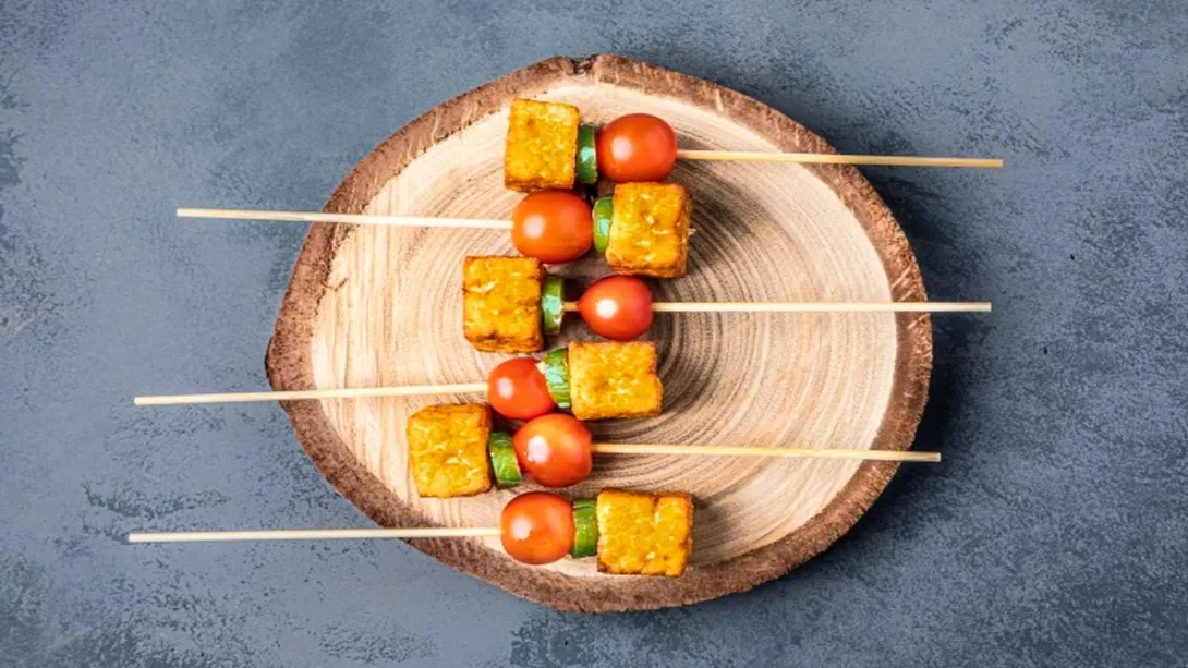 Brochetas vegetarianas con dados de tempeh, tomates cherry y pepino sobre una tabla de madera rústica. 