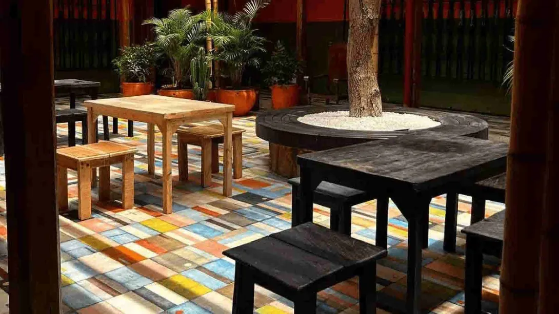 Patio interior con suelo de baldosas de colores, mesas y bancos de madera rústica, y un árbol central rodeado de piedras blancas.