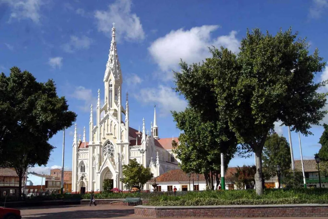 General view of the Gothic-style church with its spires reaching towards the sky and its cream-colored facade. A park with trees surrounds it