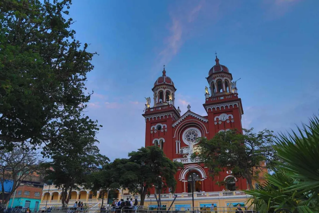 Fachada de la iglesia pintada de rojo y en un bello atardecer.  Sobresalen sus dos torres grecorromanas en forma de arcos y con figuras de ángeles a los costados.