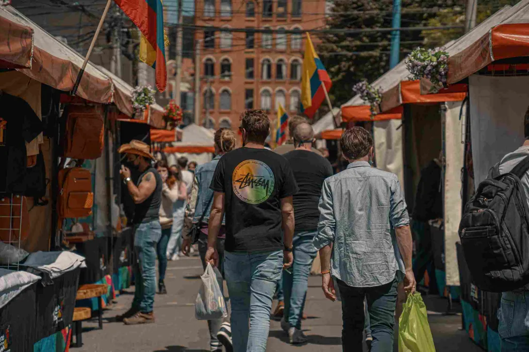  Un grupo de personas camina por entre una calle rodeada por toldos de comercio. Alrededor hay varias banderas de Colombia.