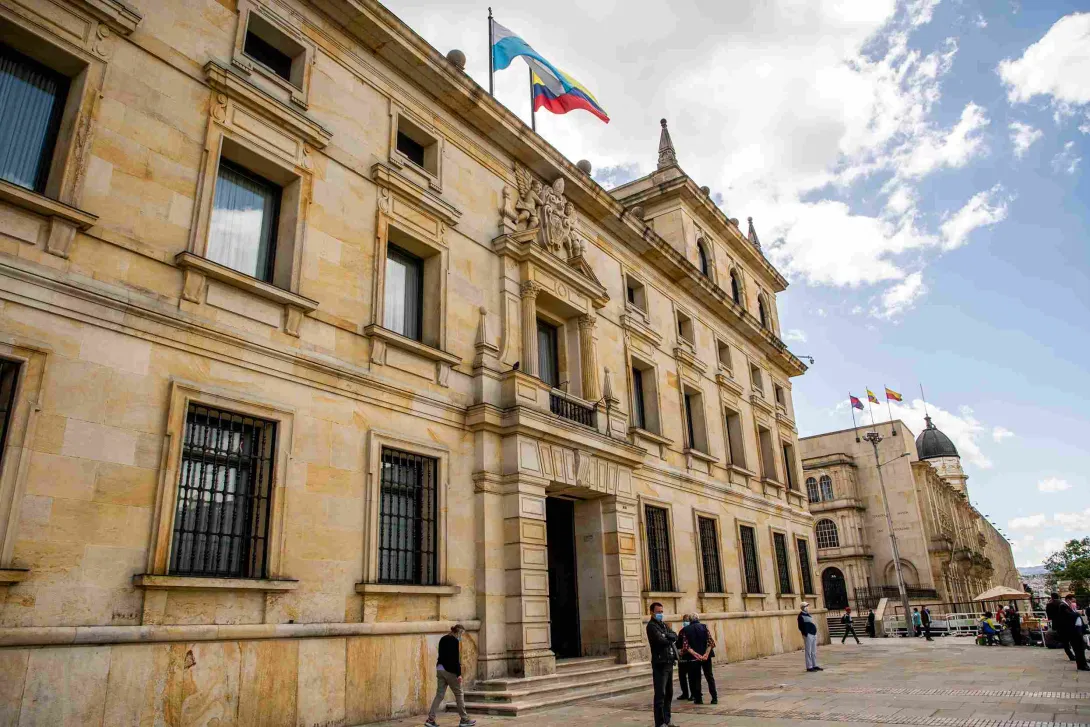  Fachada del edificio en piedra visto de lado y en el que sobresale una bandera de Colombia y otra de otro lugar, en el techo. Hay varias personas alrededor. 