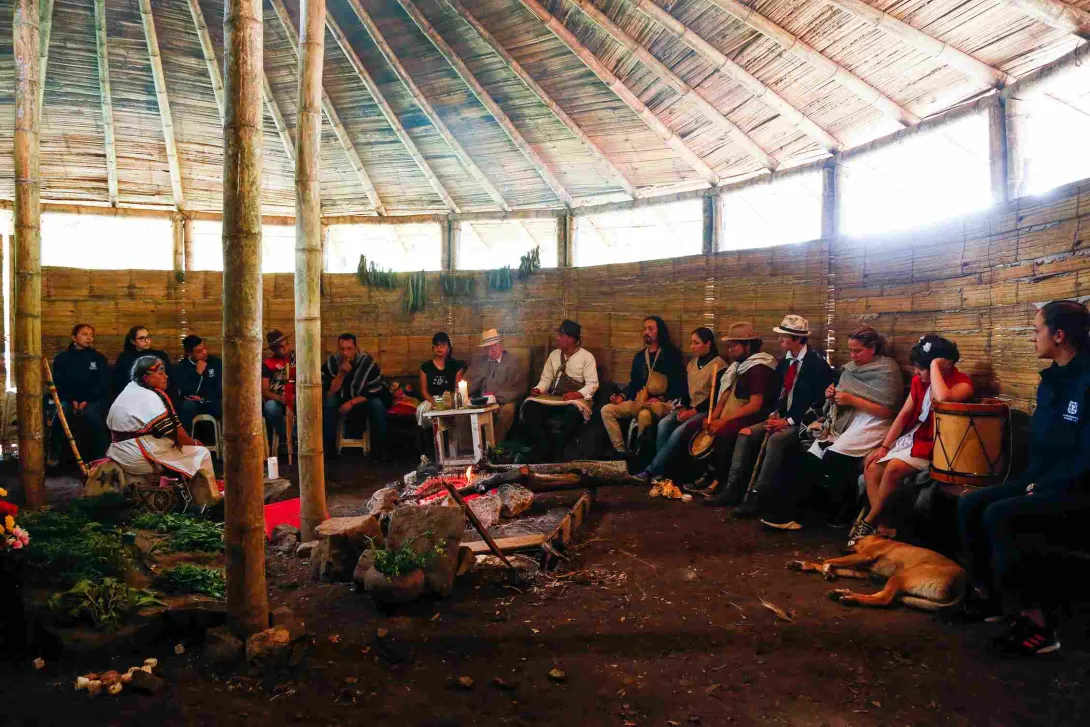 Un grupo diverso de personas reunidas dentro de una estructura circular que parece una choza tradicional. El techo está hecho de madera y paja, sostenido por pilares de madera. Las paredes están hechas de bambú y hay ventanas rectangulares a lo largo de la parte superior de la pared que dejan entrar la luz natural.