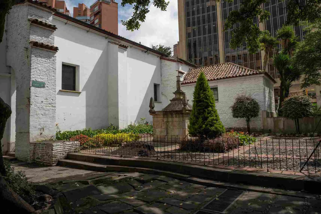 Parte exterior de la iglesia pintada de blanco. Hay un jardín con un pino y una estructura en piedra.