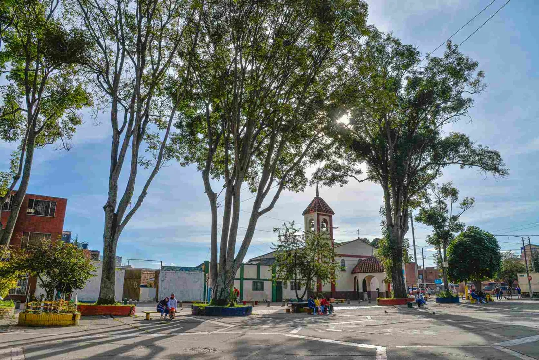 Una plaza tranquila de barrio, rodeada de altos árboles que filtran la luz del sol y proyectan sombras sobre el suelo. En el centro se alza una pequeña iglesia de fachada blanca y campanario con tejas rojas, que aporta un aire tradicional al entorno.