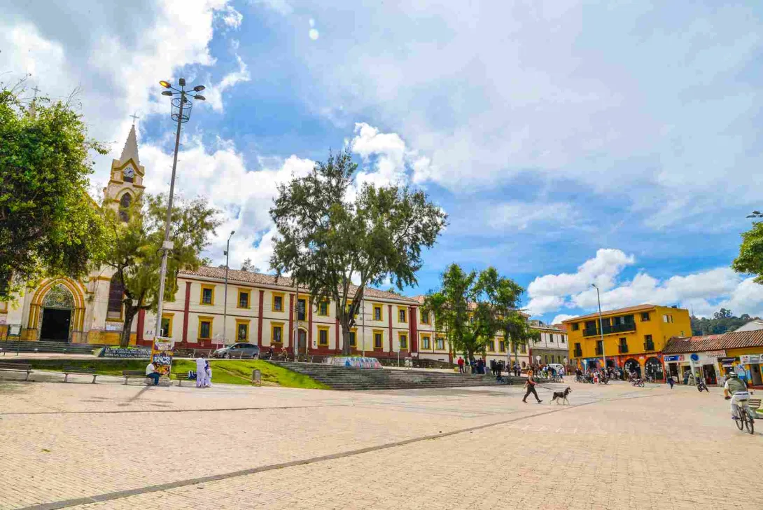 Una plaza de pueblo con una iglesia, edificios coloridos y personas disfrutando del día. En la imagen, se ve una amplia plaza pavimentada con adoquines de color claro. En el fondo, se encuentra una iglesia con una torre alta y puntiaguda, pintada de blanco y amarillo. A la derecha de la iglesia, hay un edificio de dos pisos con paredes blancas y detalles en rojo y amarillo alrededor de las ventanas. Un gran árbol está frente al edificio, proporcionando sombra.