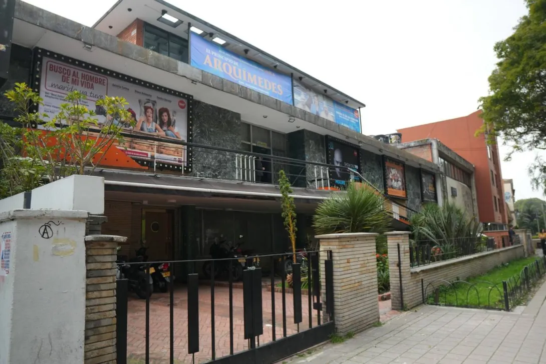 Fachada de un edificio cultural o teatro en una zona urbana, con carteles publicitarios de obras en la parte superior. La construcción tiene dos niveles, paredes de ladrillo y piedra, y un acceso con reja metálica negra. En el frente se observa un pequeño jardín con plantas ornamentales y un andén arbolado a la derecha.