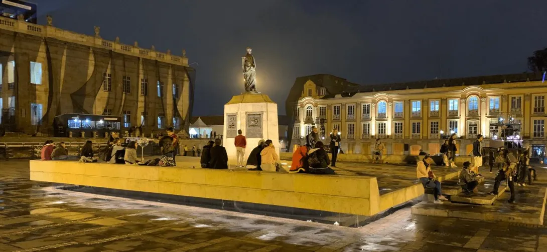 Plaza pública en horario nocturno con una estatua iluminada en el centro, rodeada de personas sentadas y conversando sobre una plataforma. Los edificios históricos alrededor están iluminados, destacando su arquitectura colonial. El suelo húmedo refleja las luces cálidas, creando un ambiente urbano animado y acogedor durante la noche.