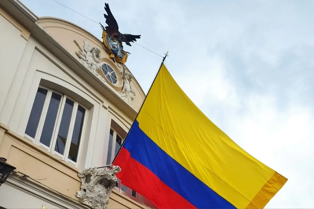  Fachada de un edificio histórico con relieves decorativos y una escultura de un condor en la parte superior, junto a una gran bandera de Colombia ondeando frente al cielo.