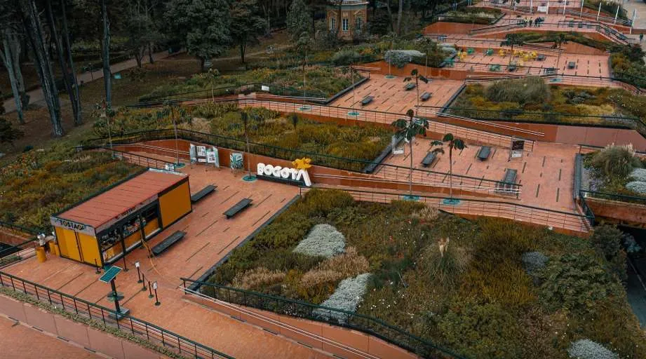 Vista aérea de un parque urbano con senderos amplios en tonos rojizos, rodeados de jardines y vegetación variada. Se observan bancas, barandas y terrazas escalonadas, además de un pequeño módulo o quiosco y un letrero que dice “Bogotá”. El entorno es verde y organizado, con áreas para el descanso y el recorrido peatonal.