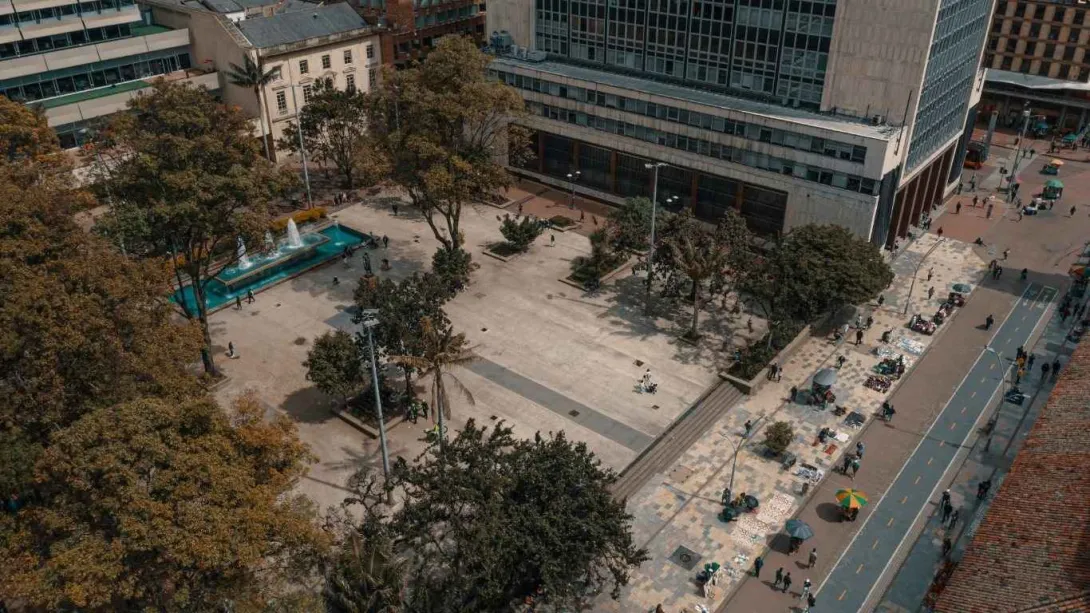 Vista aérea de una plaza urbana con amplias zonas peatonales, rodeada de edificios institucionales. En el centro hay áreas abiertas con árboles y una fuente rectangular con agua, mientras varias personas caminan o descansan en el lugar.