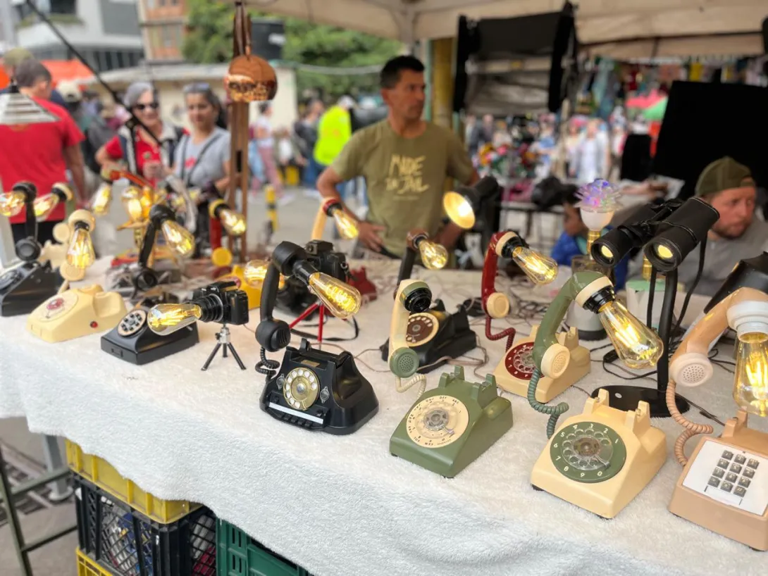  Puesto de mercado al aire libre con una mesa cubierta donde se exhiben teléfonos antiguos de disco convertidos en lámparas, con bombillas encendidas. Los objetos están organizados en filas y muestran distintos colores y diseños vintage.