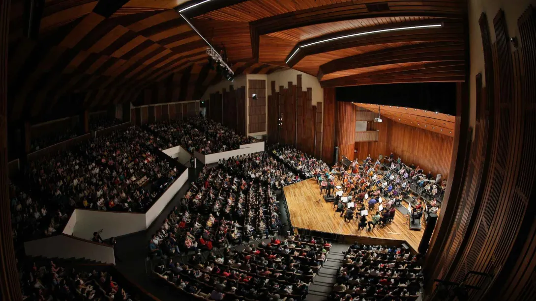 Vista panorámica del interior de un auditorio lleno de público durante un concierto. En el escenario de madera se presenta una orquesta sinfónica con numerosos músicos e instrumentos, mientras el público ocupa las graderías y platea del recinto bajo un techo curvo de madera con iluminación moderna.