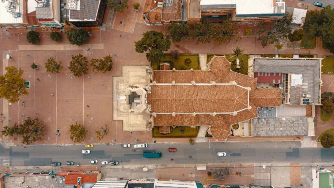 Vista aérea cenital de una iglesia con techo de tejas rojas, rodeada por una amplia plaza peatonal con árboles dispersos. A un lado se observa una vía con tráfico de carros y, alrededor, edificaciones urbanas. La composición muestra la organización del espacio público y la arquitectura del sector desde una perspectiva superior.