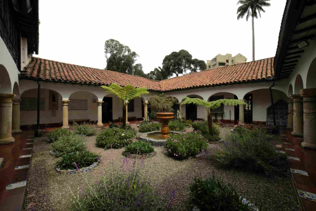 Patio interior de arquitectura colonial con corredores de arcos y columnas, techos de teja roja y un jardín central con plantas, helechos y una pequeña fuente de piedra en el centro.