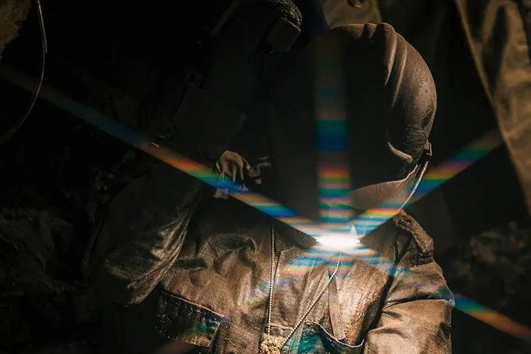 Primer plano de un trabajador utilizando una herramienta para pulir o trabajar una piedra, iluminado por una luz intensa que genera destellos de colores. La persona lleva protección en la cabeza y ropa de trabajo, en un ambiente oscuro que resalta el brillo de la luz sobre sus manos y la herramienta.