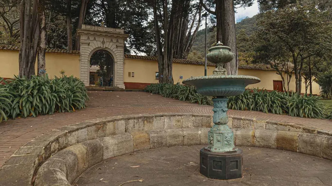 Vista del patio de la Quinta de Bolívar, donde se observa una fuente de hierro en primer plano rodeada por un pequeño estanque de piedra. Al fondo aparece el arco de entrada colonial y un camino de ladrillo flanqueado por jardines y altos árboles, con las montañas verdes de fondo.