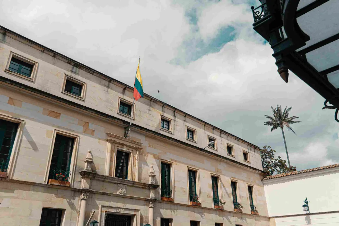 Fachada de un edificio histórico de estilo colonial con muros en tonos claros y ventanas alineadas con marcos oscuros y balcones con flores. En el centro ondea la bandera de Colombia sobre la entrada principal. El cielo está parcialmente nublado y a un costado se eleva una palma, mientras un elemento arquitectónico decorativo aparece en primer plano.